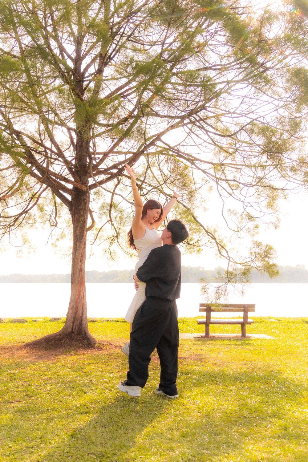 Romantic couple shares a tender moment under a large tree by a lake during golden hour.