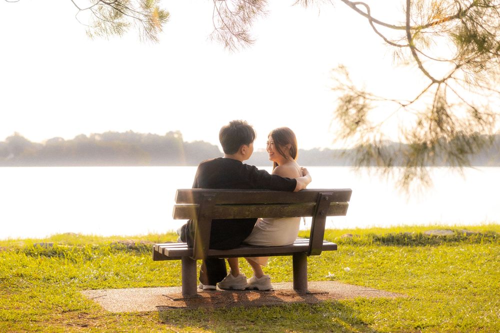 A couple sits together on a park bench overlooking a serene lake during golden hour.