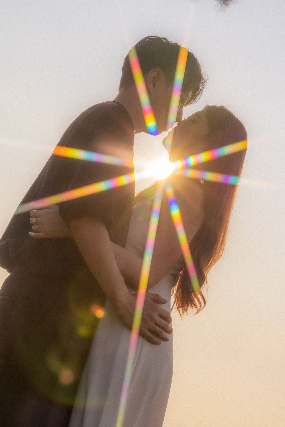 Silhouetted couple embracing with rainbow lens flare effect during sunset.
