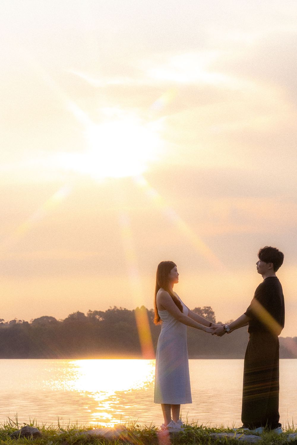 Couple holds hands at sunset by a serene lake in romantic lighting.