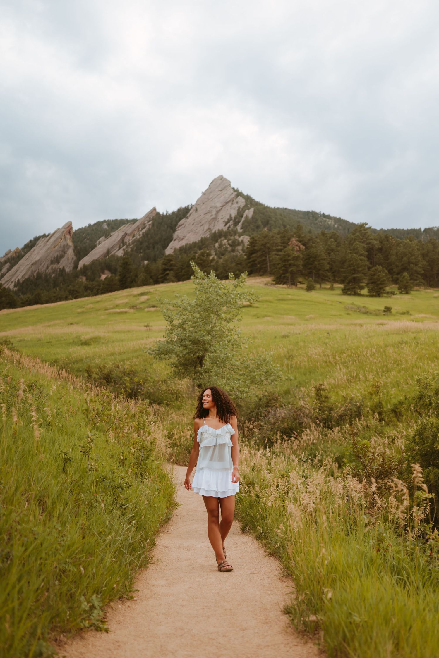 A winding dirt trail cuts through green meadows with majestic Flatirons mountains rising in the background near Boulder, Colorado.