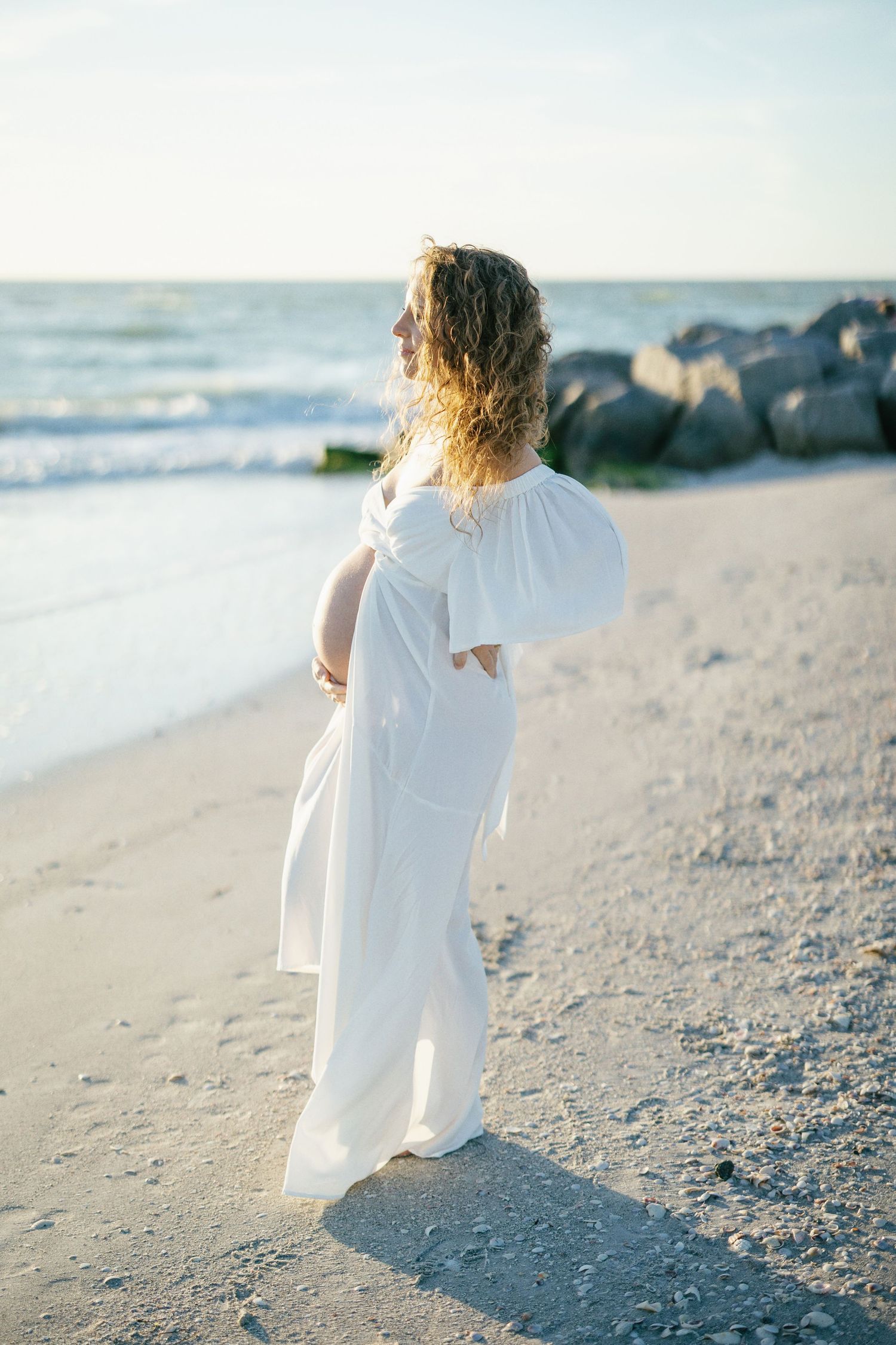 A pregnant mother in a flowing white dress stands on a sandy beach at sunset with rocky shore and ocean waves in background.
