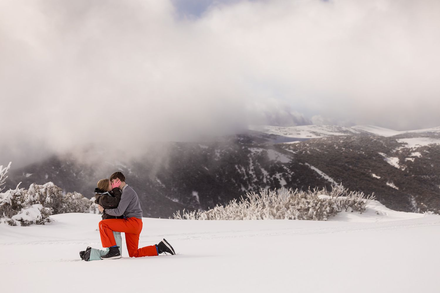 A couple share a kiss in the snow, with snow  covered mountains in the distance with mountains and trees in stunning winter landscape.