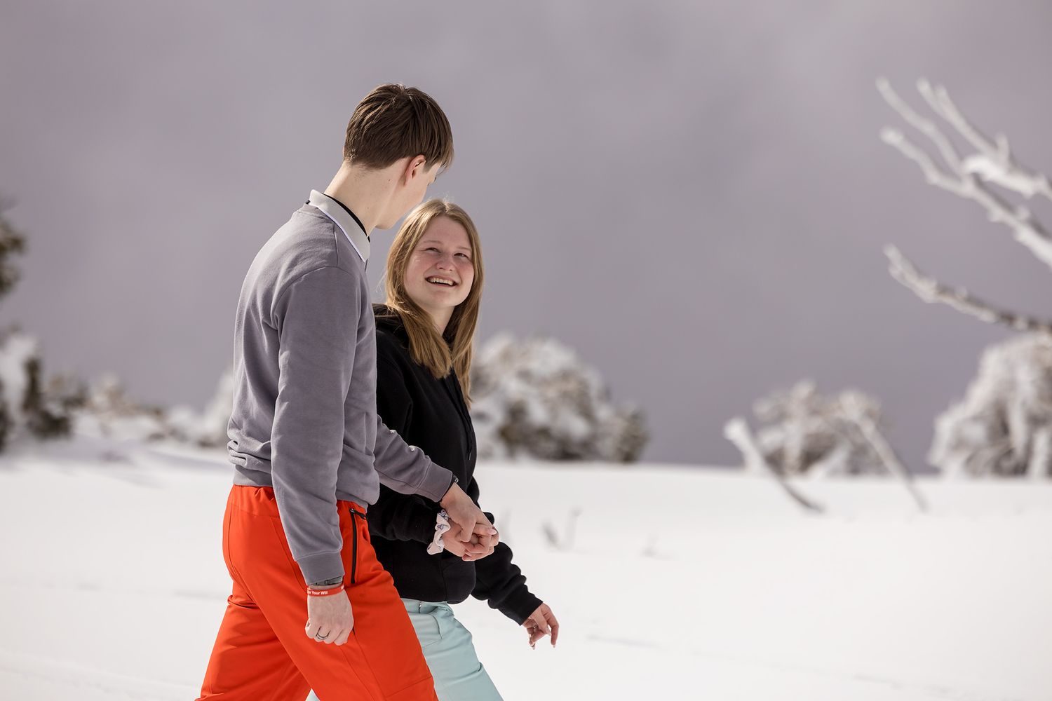 A newly Engaged couple walking holding hands on a snowy mountain slope during a winter day with cloudy skies at Falls Creek.