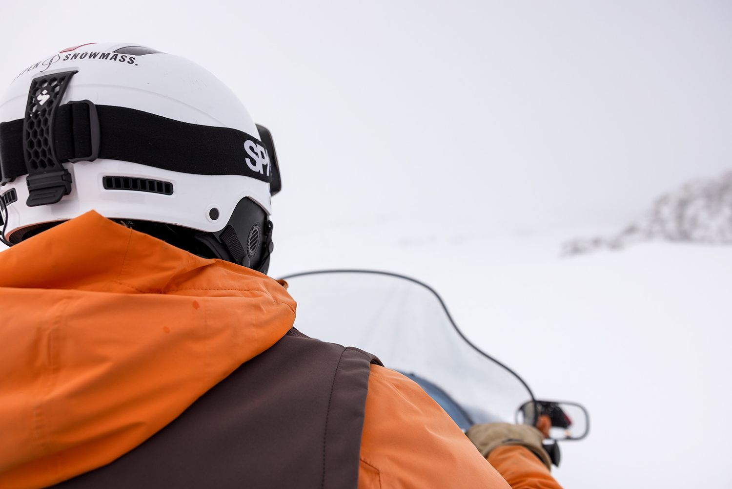 Snowmobiler wearing orange jacket and white helmet looks ahead on snowy winter terrain.