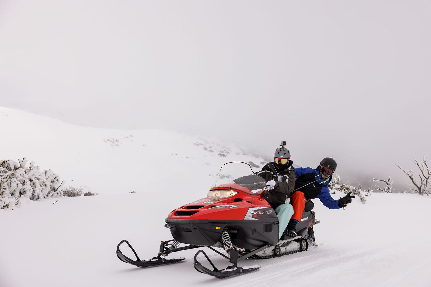 People riding snowmobiles through a snowy winter landscape with mountains visible through the fog in the background at Falls Creek.