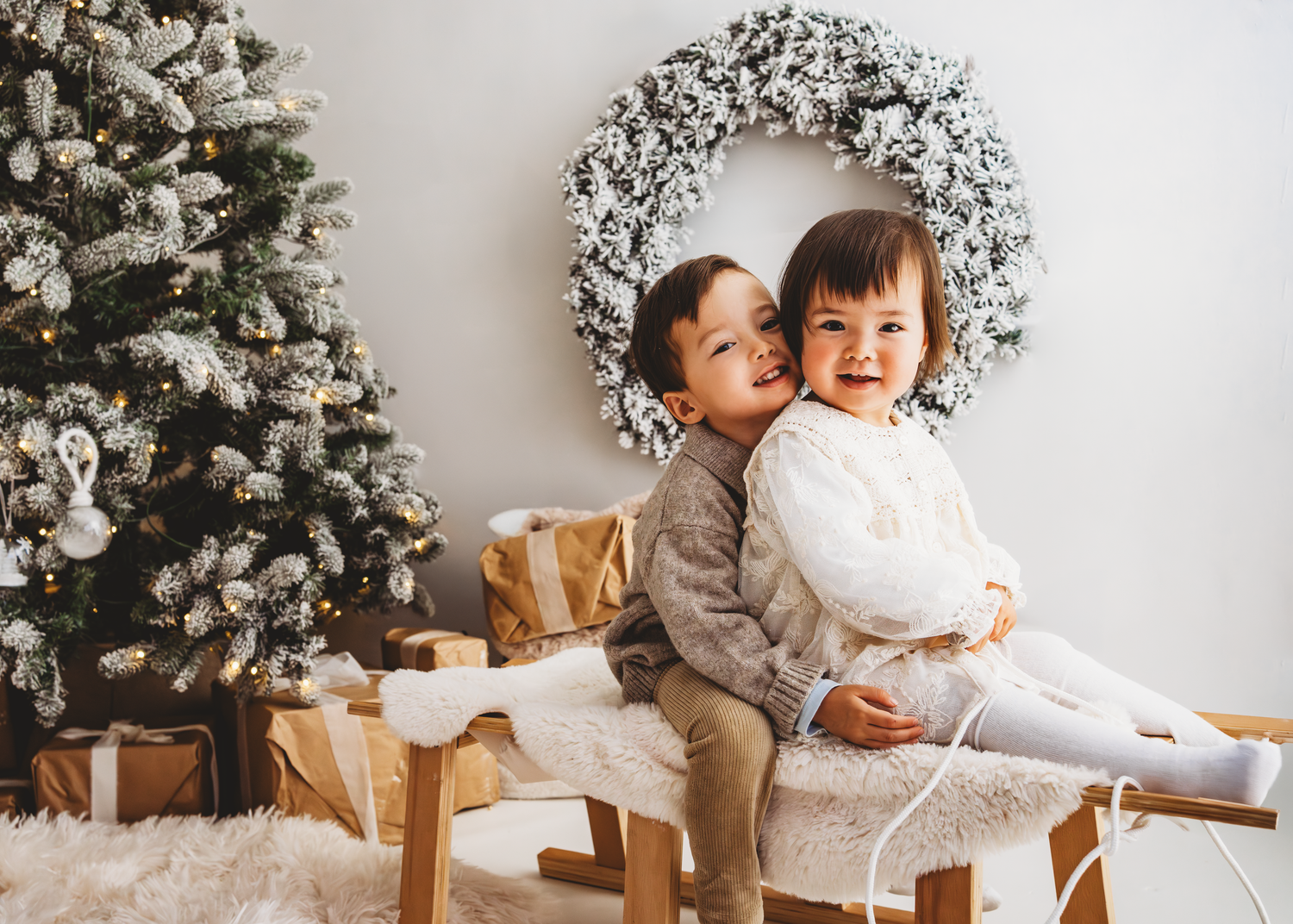 Two children in red velvet holiday dresses sit together on white bench sharing a wrapped present by a snowy wreath. Christmas Mini Photoshoo