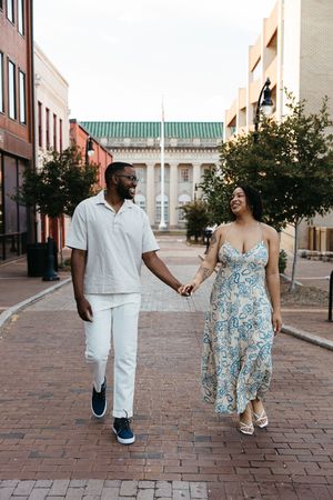 Two people hold hands while walking together on brick pavement near classic architecture and trees.