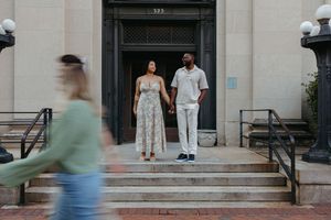 A couple dressed in white attire stands at the entrance of a historic building while a blurred figure walks past.