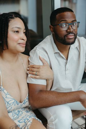 A loving couple sits close together, with the woman wearing a floral dress and the man in glasses and a white shirt.