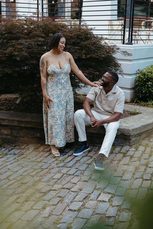 A couple sits together on stone steps, with one wearing a floral maxi dress and another in casual white attire.