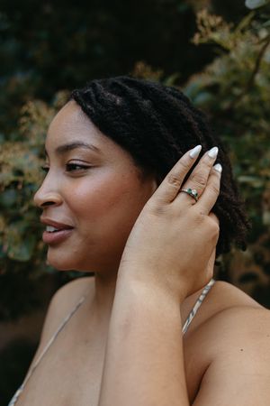 Profile view of someone wearing a ring and long nails, shown against a natural outdoor background.