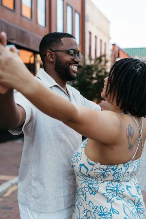 Romantic couple in coordinated white and blue outfits dancing outdoors on a warm evening by brick buildings.