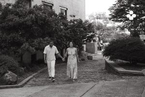 A vintage black and white photograph shows a couple walking down a tree-lined residential street in summer attire.
