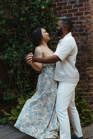 A couple embraces and dances together against a brick wall with lush greenery, wearing white and a blue floral dress.