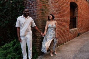 A couple dressed in elegant casual attire walk hand in hand along a rustic brick wall pathway.