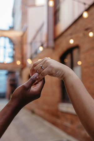 Two hands reaching towards each other to touch against a backdrop of brick walls and string lights at dusk.