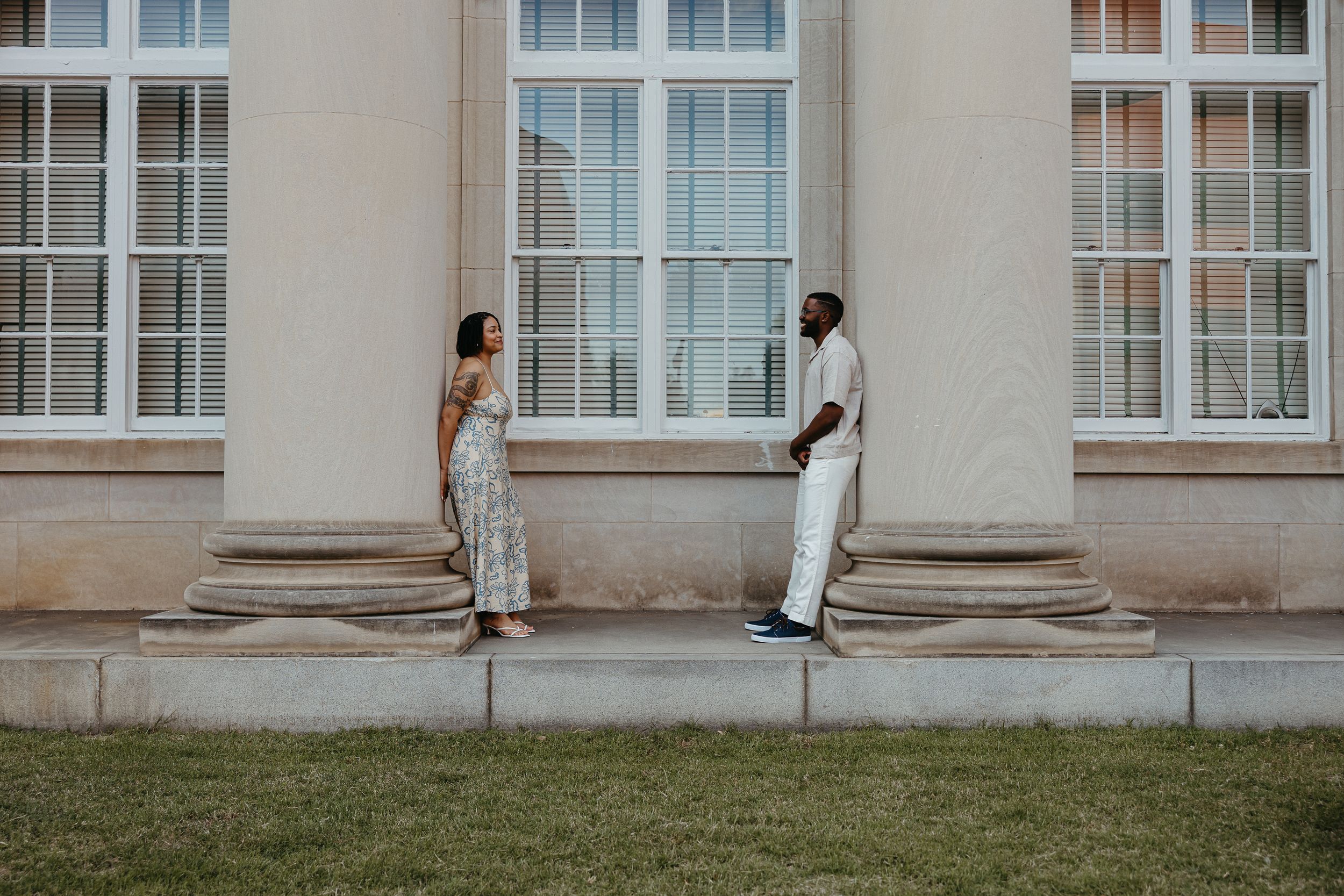 Two people in elegant white outfits lean against white pillars of a classical building, creating a symmetrical composition.
