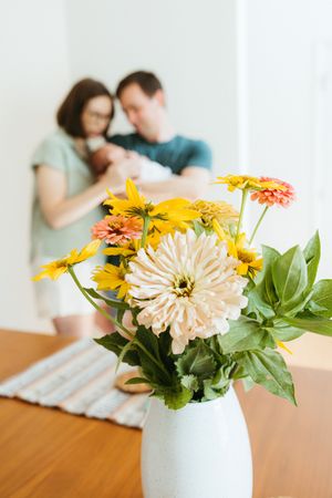 Fresh bouquet of yellow and cream flowers arranged in a white vase atop a wooden dining table.