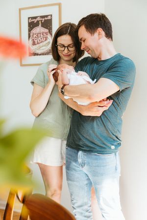 Two people standing together, cradling a newborn baby at home, with a vintage-style poster on the wall behind them.