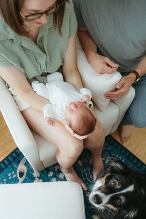 Newborn baby rests on white couch with someone wearing sage green shirt while black and white dog looks on.