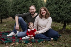 Happy family of three poses on plaid blanket surrounded by evergreen trees.