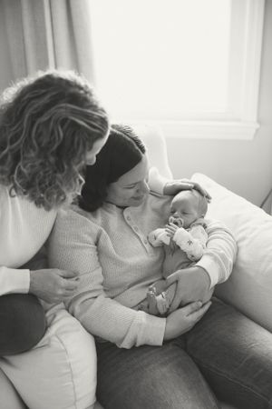 A tender black and white photograph captures an intimate family moment with a sleeping newborn on a cozy couch at home.