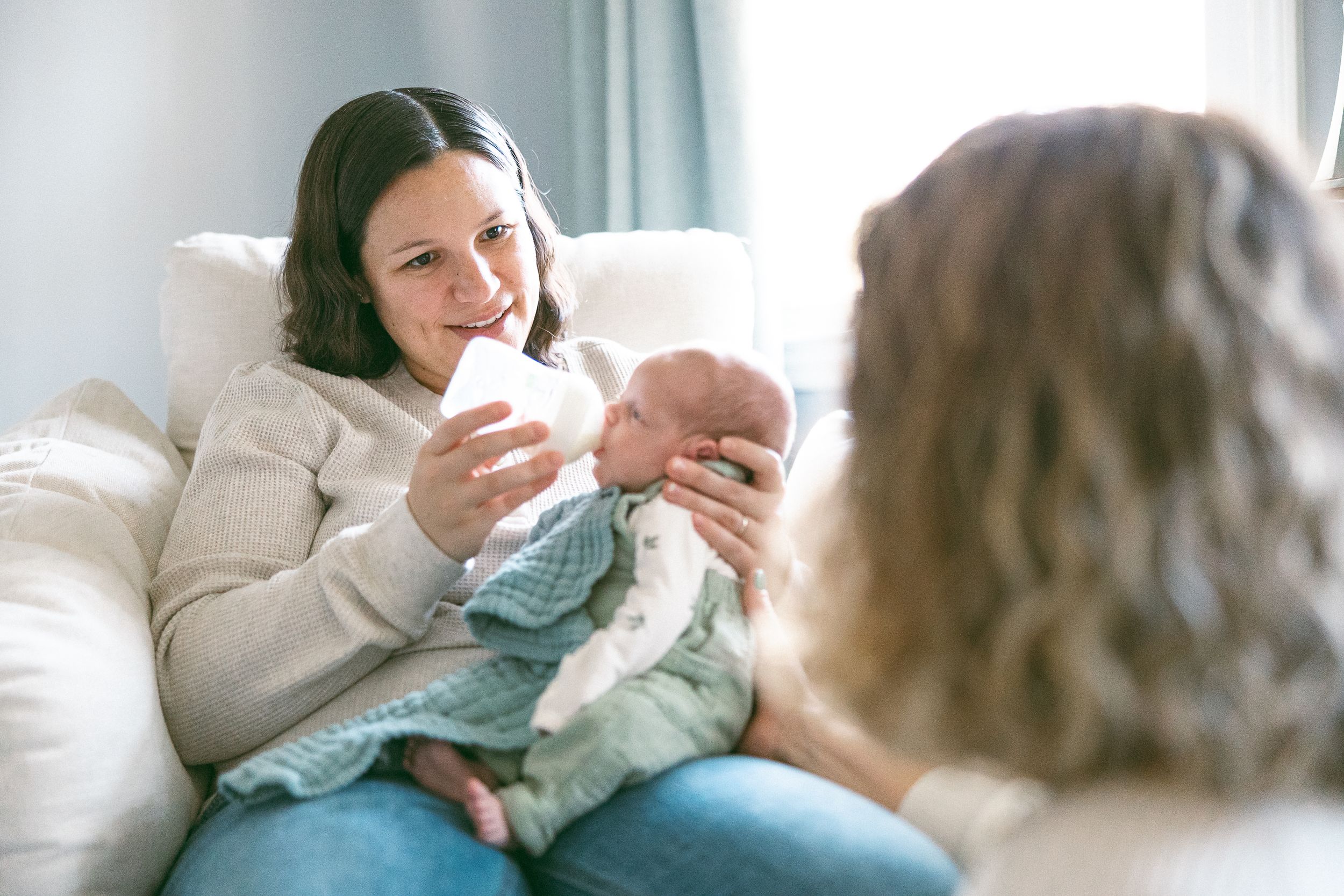 In a softly lit room, a mother in a cream sweater sits on a couch bottle-feeding her baby with another person nearby.