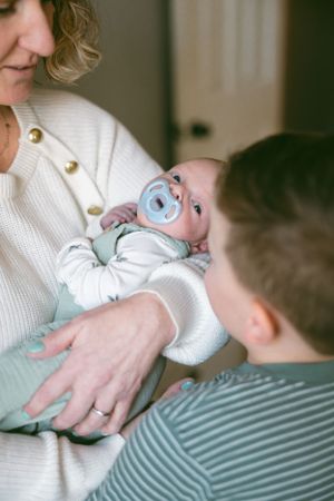 A baby wearing blue with a pacifier is cradled while being lovingly watched by family members.