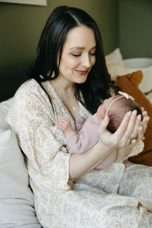 New mother in lace robe holds tiny sleeping newborn baby while sitting on bed in soft natural light.