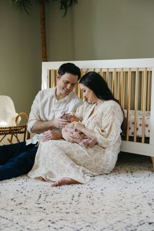 Parents lovingly cradle their newborn baby while sitting together on a carpeted floor beside a white crib.