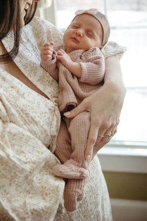 A newborn baby wearing a soft pink knitted outfit smiles contentedly while being cradled in loving arms by a window.