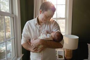 A tender moment by a sunlit window as a parent cradles a sleeping newborn baby wearing a white onesie.