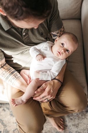 A tender parental moment captured as baby in white onesie lies on caregiver's lap on a beige couch.
