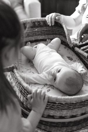 A baby lying in a woven basket wearing white clothing while hands reach to touch them in black and white.