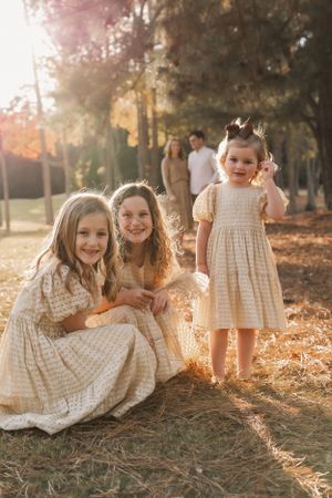 Three young children in vintage-style cream lace dresses pose together in a sunlit forest setting during golden hour.