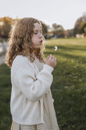 A person with curly hair wearing a white knit sweater blows on a dandelion in a sunlit field during golden hour.
