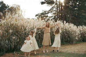 Three people in flowing dresses dance together in a golden field of pampas grass at sunset.