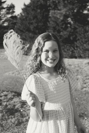 Black and white vintage photo of someone in a white dress holding a tall grass stalk outdoors on a sunny day.