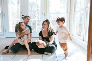 Family of five enjoying a relaxed moment together on a sunlit tiled floor near large windows at home.