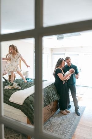 Family in bedroom with parents holding newborn baby while young girl jumps on bed behind them.