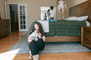 A mother sits on a bedroom floor nursing a newborn while two children play on the bed behind her.