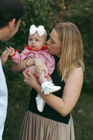 A baby in a red and white dress is held and interacted with during an outdoor photo sequence.