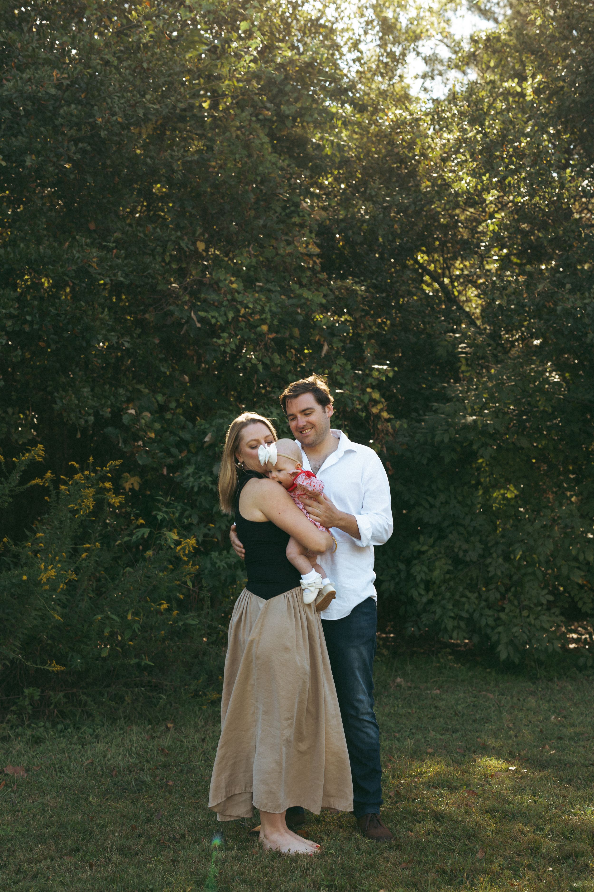 A couple stands barefoot on grass holding a baby while surrounded by lush green trees in golden sunlight.