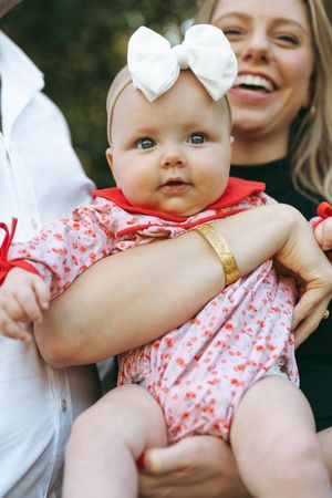 Baby wearing a white bow and red floral romper is held while smiling outdoors.