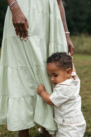 A small child in white clothing holds onto a flowing sage green dress in an outdoor setting.
