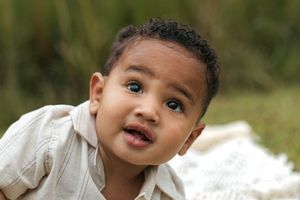 A small child in a white outfit smiles while sitting outdoors in a natural setting.