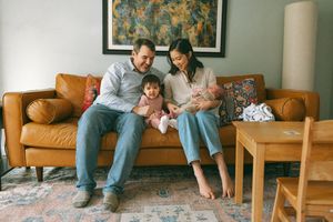 A family sits together on a tan leather sofa in their living room with wooden furniture and a patterned rug.