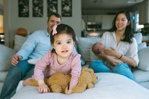 A family relaxes together on a white couch at home with a child holding a teddy bear.