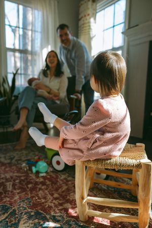 Small child sits on wooden stool in sunlit room while parents relax in background on comfortable sofa.